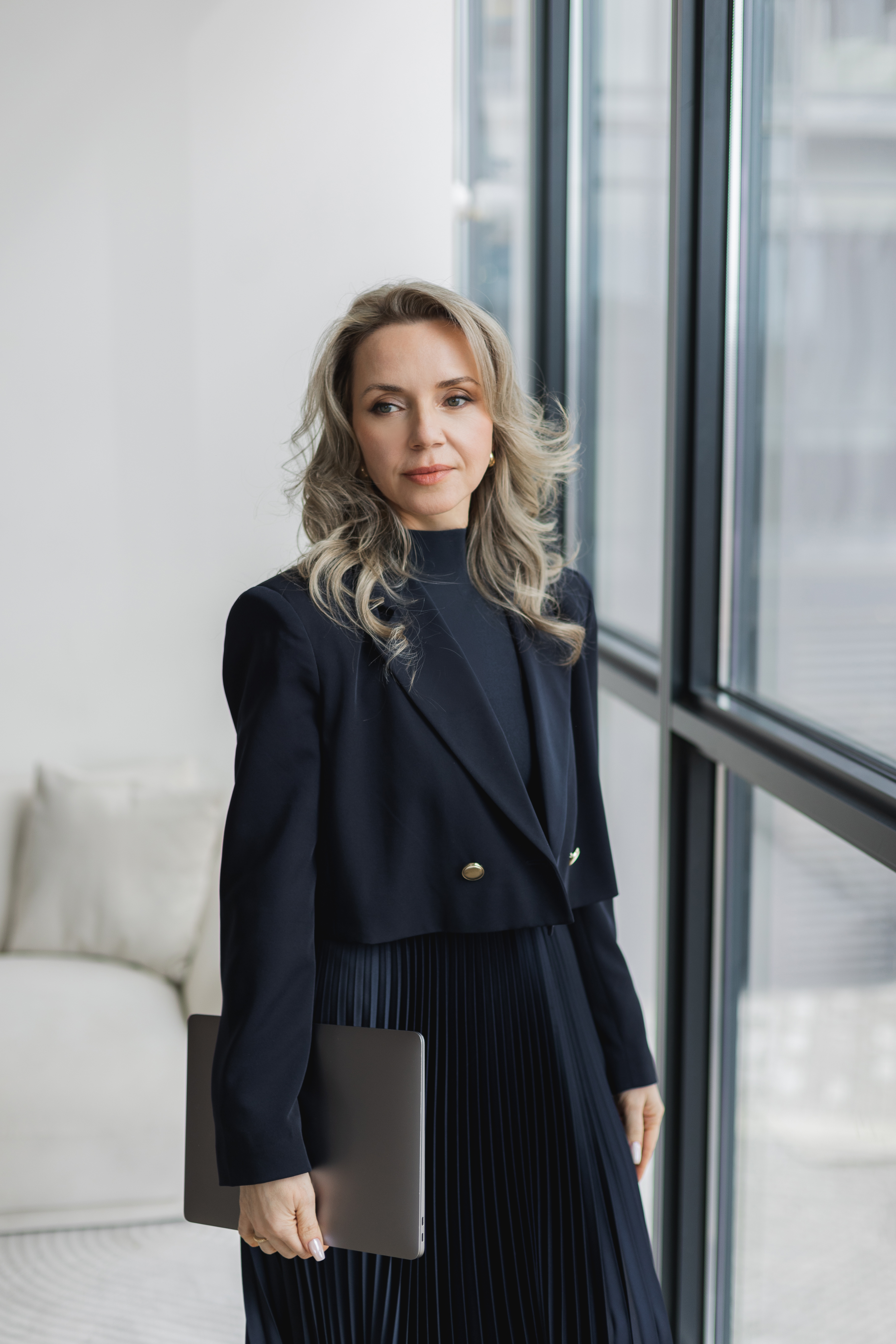 Yana Mamotenko in a navy blazer holding a laptop by a floor-to-ceiling window